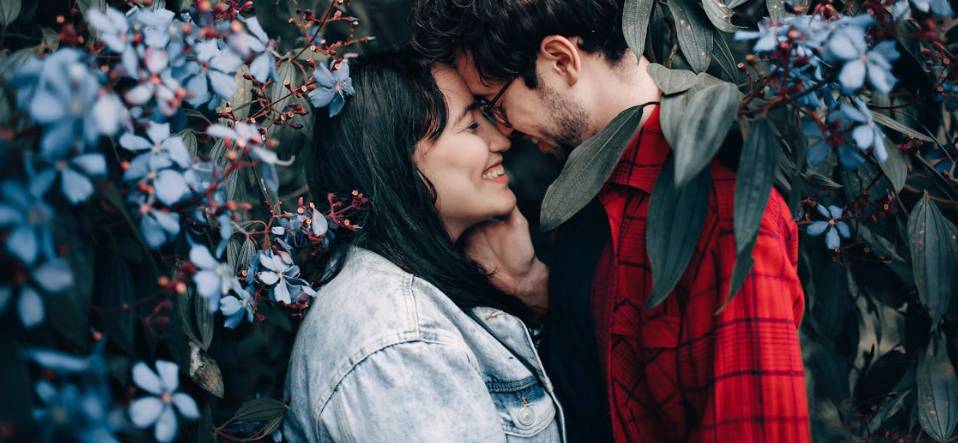 Casal sorrindo em clima romântico entre flores roxas em cenário natural e intimista