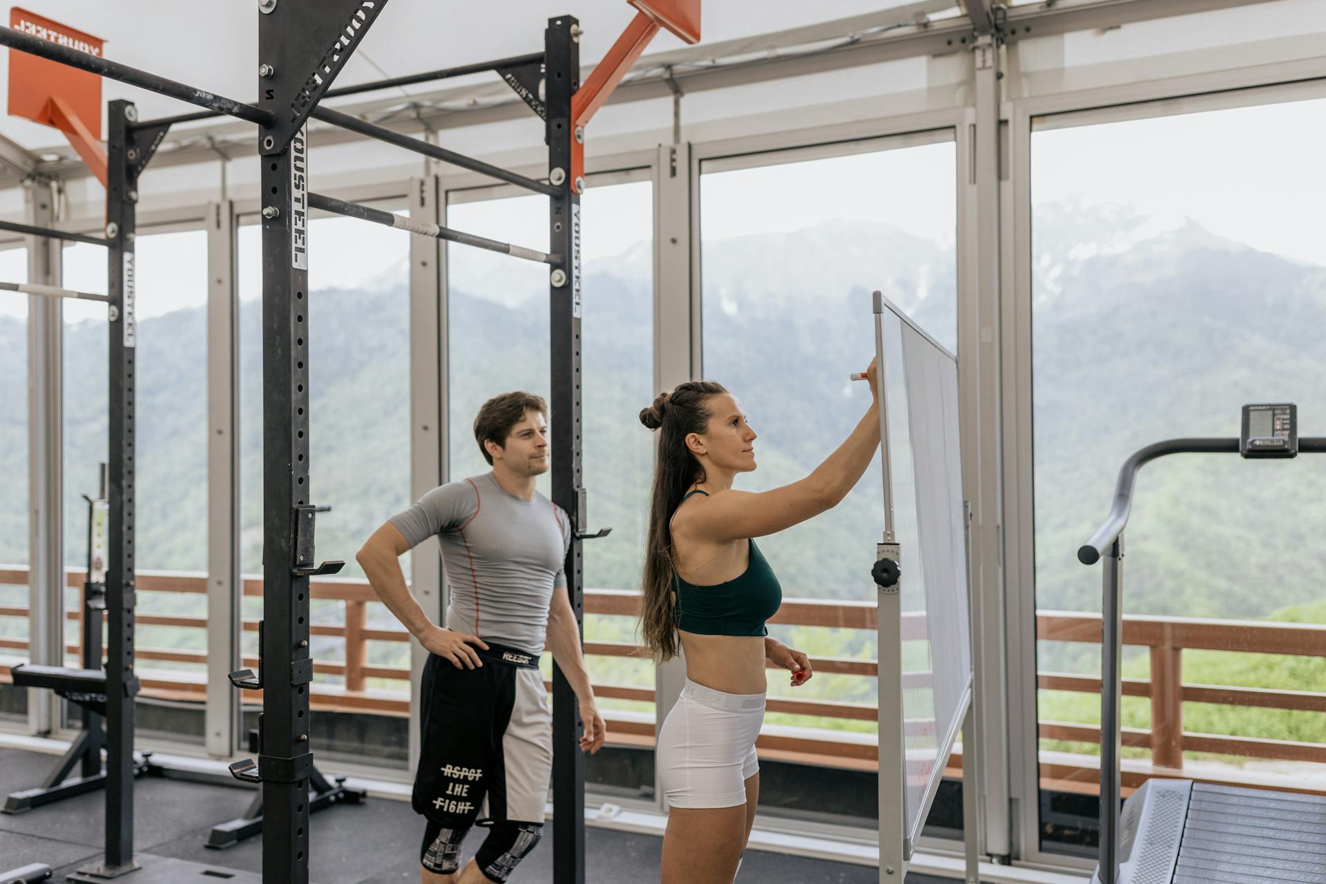 Personal trainer anotando plano de treino em quadro enquanto aluno observa na academia