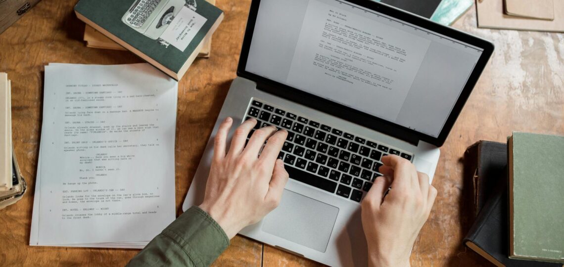 Mãos digitando texto persuasivo em notebook rodeado por livros e roteiros sobre mesa de madeira