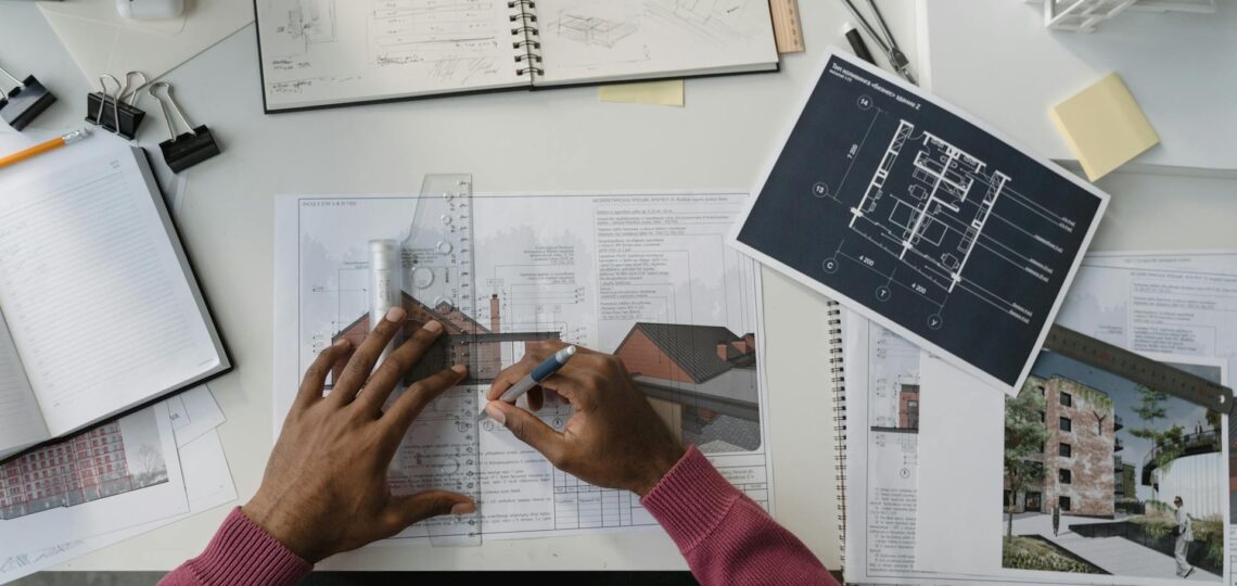 Mãos desenhando projetos arquitetônicos com plantas e maquetes sobre a mesa