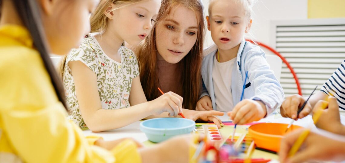 Professora orientando crianças em atividade artística com tintas em sala de aula criativa.