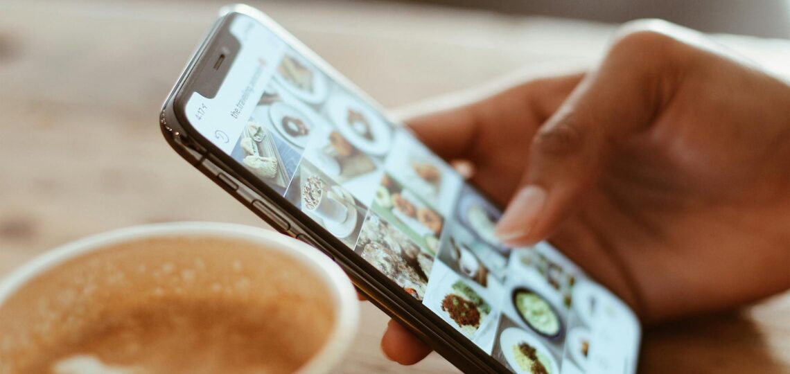 Pessoa segurando smartphone enquanto navega por posts de comida no Instagram, com xícara de café em primeiro plano.