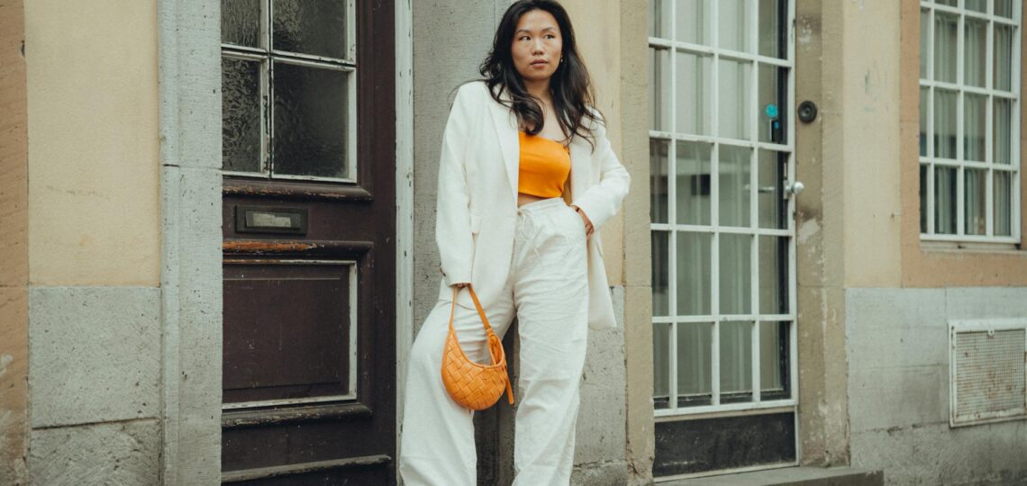 Mulher usando look moderno com blazer branco e top laranja, encostada em porta de madeira no centro urbano europeu.