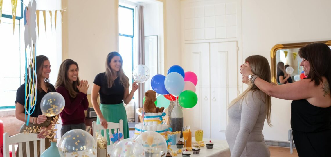 Mulheres celebrando um chá de bebê com balões, doces e decoração alegre em ambiente interno iluminado