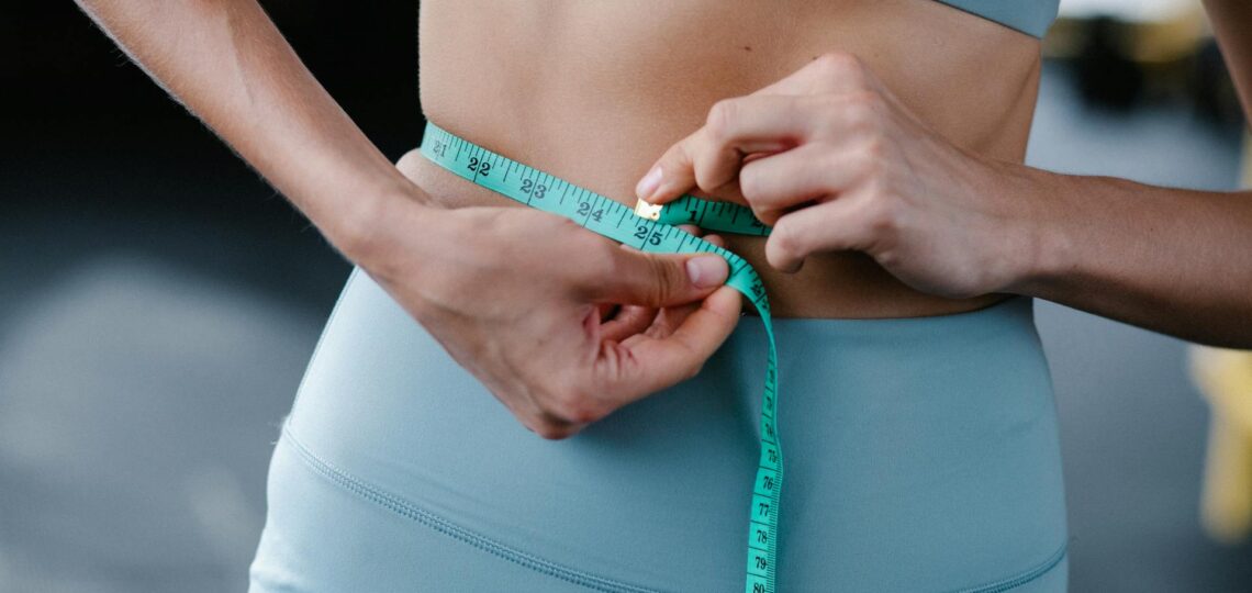 mulher com roupa de ginantica tirando medida da cintura com fita métrica