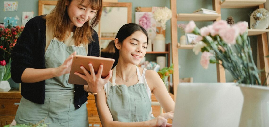 duas mulheres que trabalham em uma floricultura acessando um tablet e um laptop sobre a mesa