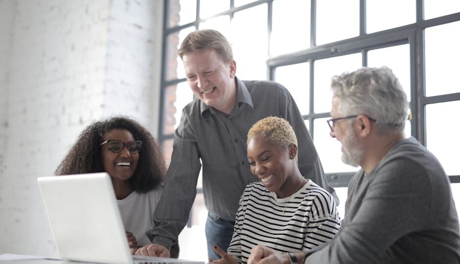 Group of diverse people laughing and using netbook while working on project together in office with white brick walls and big window