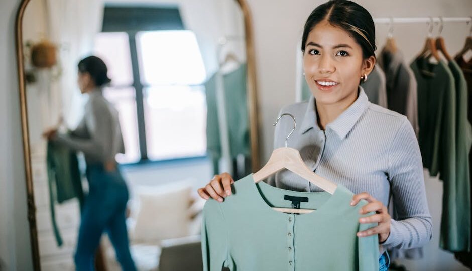 Young woman holds light green blouse in stylish indoor setting, smiling and confident.