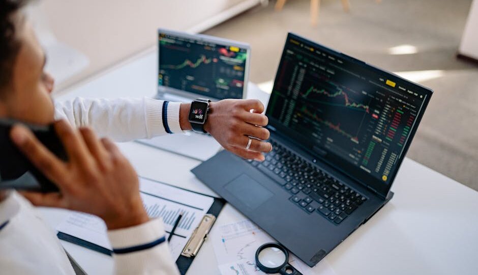 Man multitasking with a smartwatch and laptop analyzing stock market charts at the office.