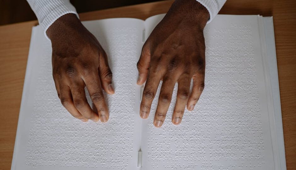 A pair of hands reading a braille book on a wooden table, emphasizing touch and sensory experience.