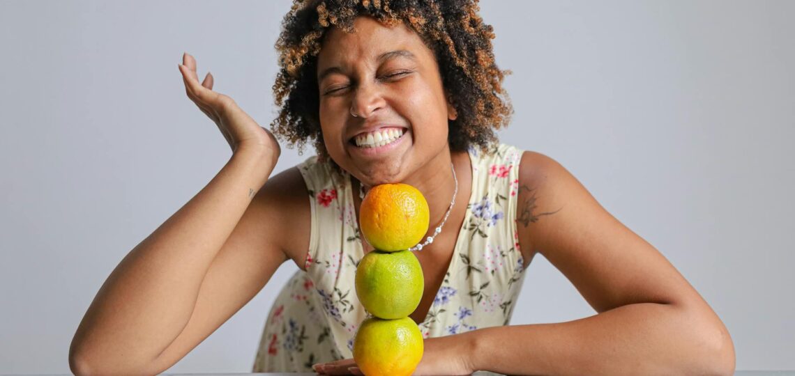 mulher de 40 anos sorrindo equilibrando 3 laranjas sobre uma mesa