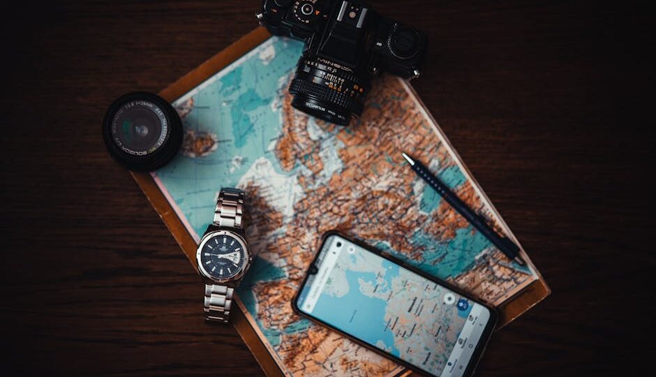 Top view of travel essentials including a map, camera, phone, watch, and lens on a wooden surface.
