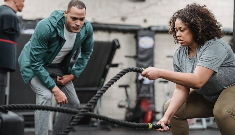 Plus sized African American female in sportswear exercising with battle ropes near serious male instructor during intense workout in modern fitness center