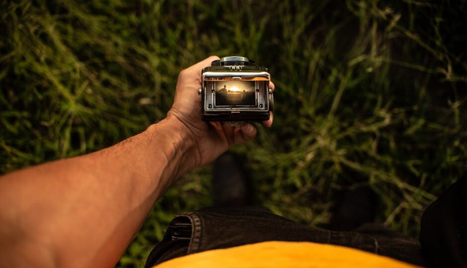 From above of faceless male traveler with twin lens photo camera with screen in hand standing on grassy ground in countryside