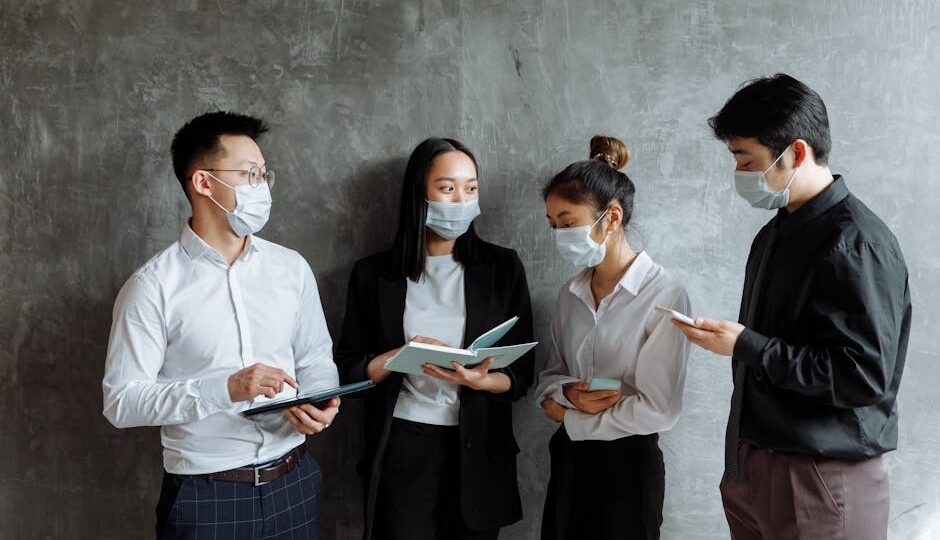 Four professionals wearing face masks collaborate with tablets and notebooks indoors.