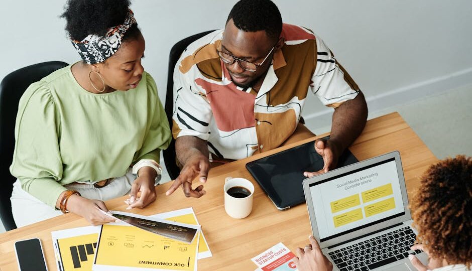 Three colleagues collaborate over marketing documents and a laptop in a modern office setting.