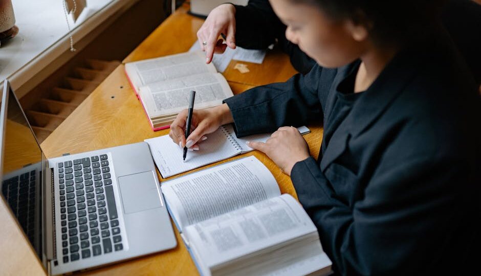 Two students engaged in study, taking notes with books and a laptop on a wooden desk indoors.