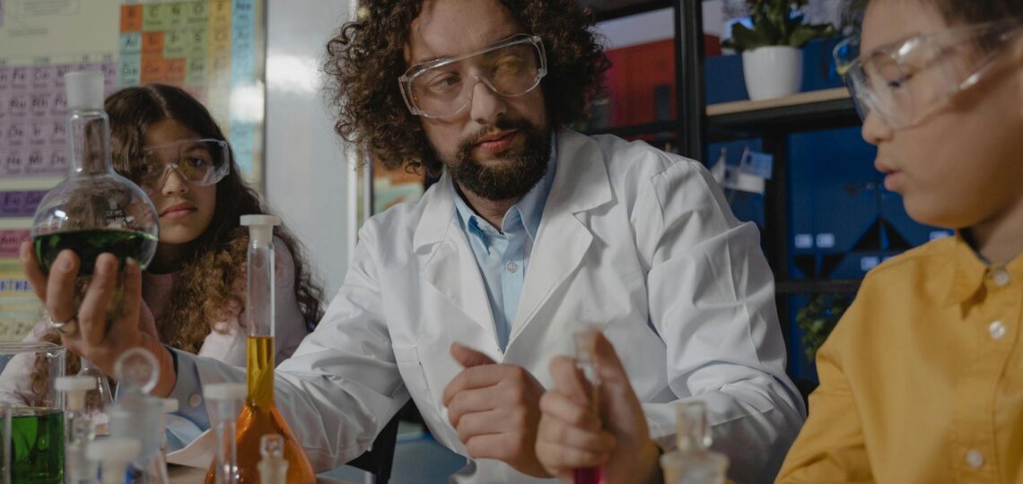 aula de ciências com professor ensinando duas jovens em frente a experimentos de ciência