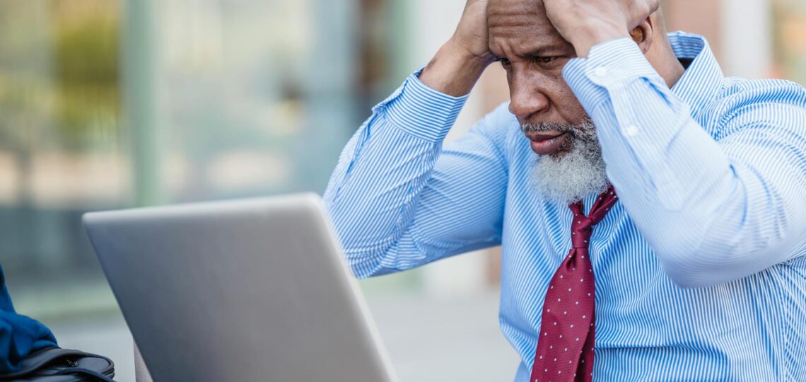 homem executivo com as mãos na cabeça desesperado olhando para a tela de um laptop